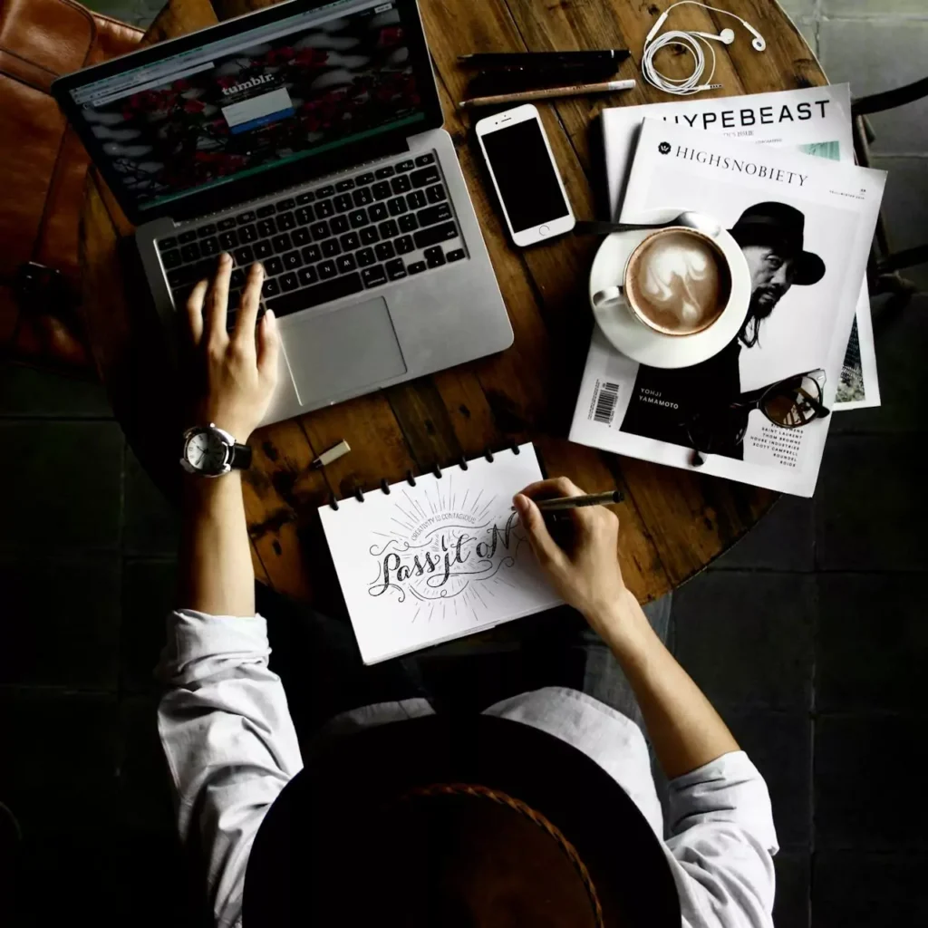 Ein Tisch mit einem Laptop, Notizbuch, Kaffeetasse und Zeitschriften. Eine Hand zeichnet.